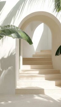 The interior showcases a minimalist design with white walls and large archways. Lush green plants frame the space, creating a serene and fresh atmosphere. The soft light enhances the tranquil setting.